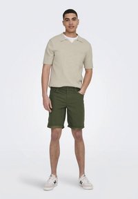 Zaļas šorti ar uzlocītām malām, kopā ar gaiši bēšu īsrokas adījuma topu. Sporta apavi ir balti ar gaiši pelēkiem akcentiem.