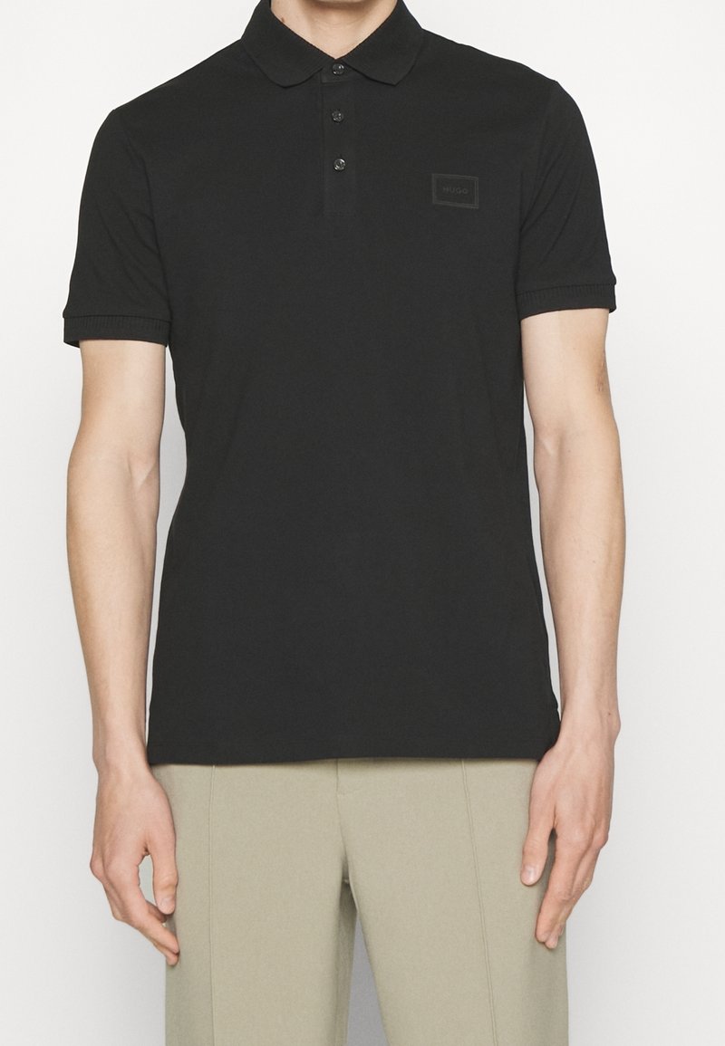 Homme portant un polo noir à manches courtes avec une patte de boutonnage à trois boutons et un pantalon beige, debout devant un fond uni.