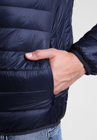 Marinblå pufferjacka med kviltad textur, med en sidoficka, justerbara ärmslut och en slät yta. Närbild av hand i fickan.