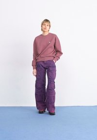 Violet spalvos cargo kelnės su keliais kišenėmis, derinamos su prigludusiu bordo spalvos džemperiu, ant baltos fono ir mėlyno grindų paviršiaus.
