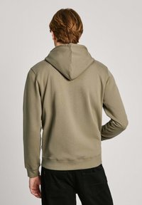 Sudadera caqui con capucha ajustable, mangas largas y puños acanalados. La tela tiene una textura suave, combinada con pantalones negros.