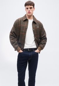 Νέος άνδρας που φοράει καφέ καρό σακάκι πάνω από λευκό πουκάμισο, σκούρα τζιν και μαύρη ζώνη, στέκεται με τα χέρια στις τσέπες.