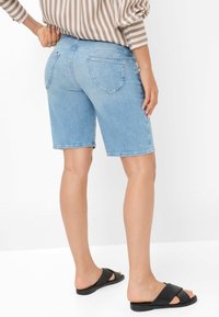 Shorts en denim bleu clair avec poches arrière, ourlet effiloché et coupe ajustée, associés à des sandales noires à brides croisées. Fond clair.