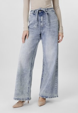 Γυναίκα που φοράει τζιν παντελόνι υψηλής μέσης, ανοιχτόχρωμο με φαρδιά πόδια, ζώνη με φιόγκο και μπεζ μυτερές γόβες μπροστά σε μονόχρωμο φόντο.