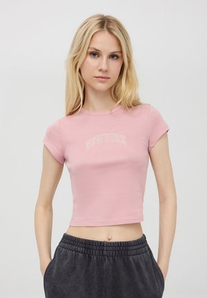 T-shirt corta rosa con maniche corte, che presenta "NEW YORK" in lettere bianche sul petto. Tessuto morbido e elastico con un design aderente.