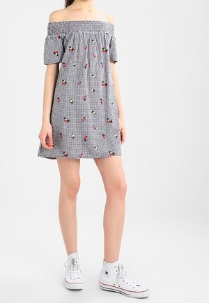 Kvinde iført off-shoulder sort-hvid ternet kjole med blomster mønstre og hvide høje sneakers, stående mod en ensfarvet baggrund.