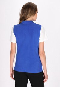 Blauer Fleece-Weste mit Kragen, die über einem weißen kurzärmeligen Hemd getragen wird. Die Weste hat eine glatte Textur und keine sichtbaren Verschlüsse.