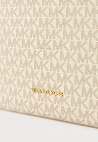 Beige handtas gemaakt van textuurmateriaal met een herhaald logo-patroon. Het heeft gouden hardware met de merknaam.