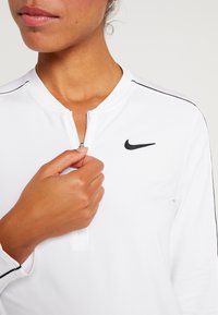 Langärmliges, weißes Nike-Oberteil mit einem halben Reißverschluss, das mit einem schwarzen Logo und seitlichen Streifendetails versehen ist. Der glatte, leichte Stoff schmiegt sich an den Körper an.