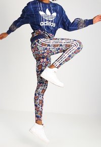Felpa blu navy con logo Adidas bianco, abbinata a leggings con motivo floreale e strisce laterali, e sneakers bianche.