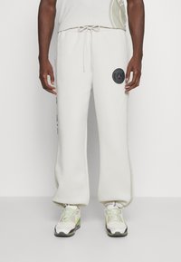 Lys beige sweatpants med snoretræk i taljen, sidelommer og et sort cirkulært logo på venstre lår. Blød tekstur og afslappet pasform.