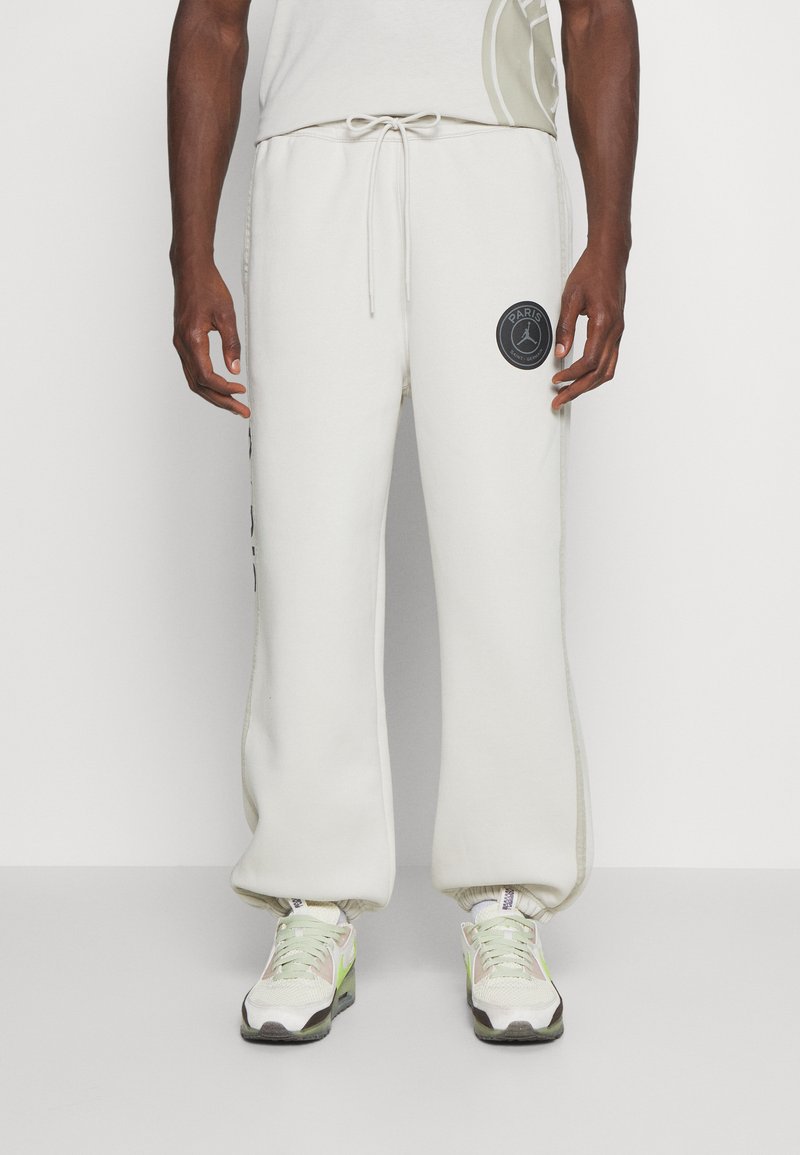 Lys beige sweatpants med snoretræk i taljen, sidelommer og et sort cirkulært logo på venstre lår. Blød tekstur og afslappet pasform.