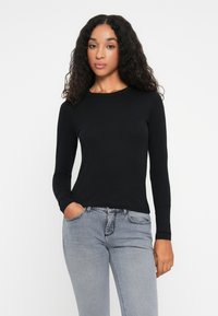 Γυναίκα με σγουρά μαλλιά, φορώντας ένα εφαρμοστό μαύρο μπλουζάκι με μακρύ μανίκι και ανοιχτό μπλε τζιν, με το ένα χέρι στην τσέπη, στέκεται σε λευκό φόντο.