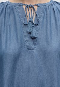 Blouse en denim bleu avec un décolleté en V et des cordons à pompons noués au col, présentant un tissu froncé aux épaules.