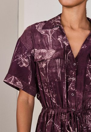 Frau trägt ein kurzärmeliges, knöpfbares lila Kleid mit Blumenmuster und elastischem Bund, der mit einer Kordel zusammengebunden ist.