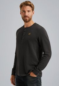 Tamno siva dug rukav henley majica s okruglim izrezom, tri dugmeta na prednjem dijelu i malim zlatnim logotipom. U kombinaciji s plavim trapericama.
