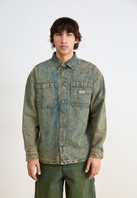 Camicia in denim blu sbiadito con due tasche frontali, chiusura a bottone e colletto. Presenta un tessuto testurizzato e una vestibilità morbida.
