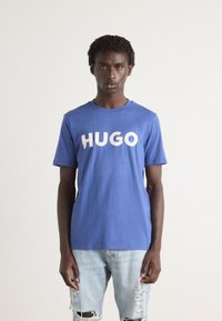 Kék pamut póló, nagy fehér "HUGO" felirattal az elején. Világoskék, kopottas farmernadrággal párosítva. Egyszerű, hétköznapi dizájn.