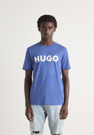 Blå bomull t-skjorte med stor hvit "HUGO" tekst på forsiden. Kombinert med lys blå slitte denimjeans. Enkel, uformell design.