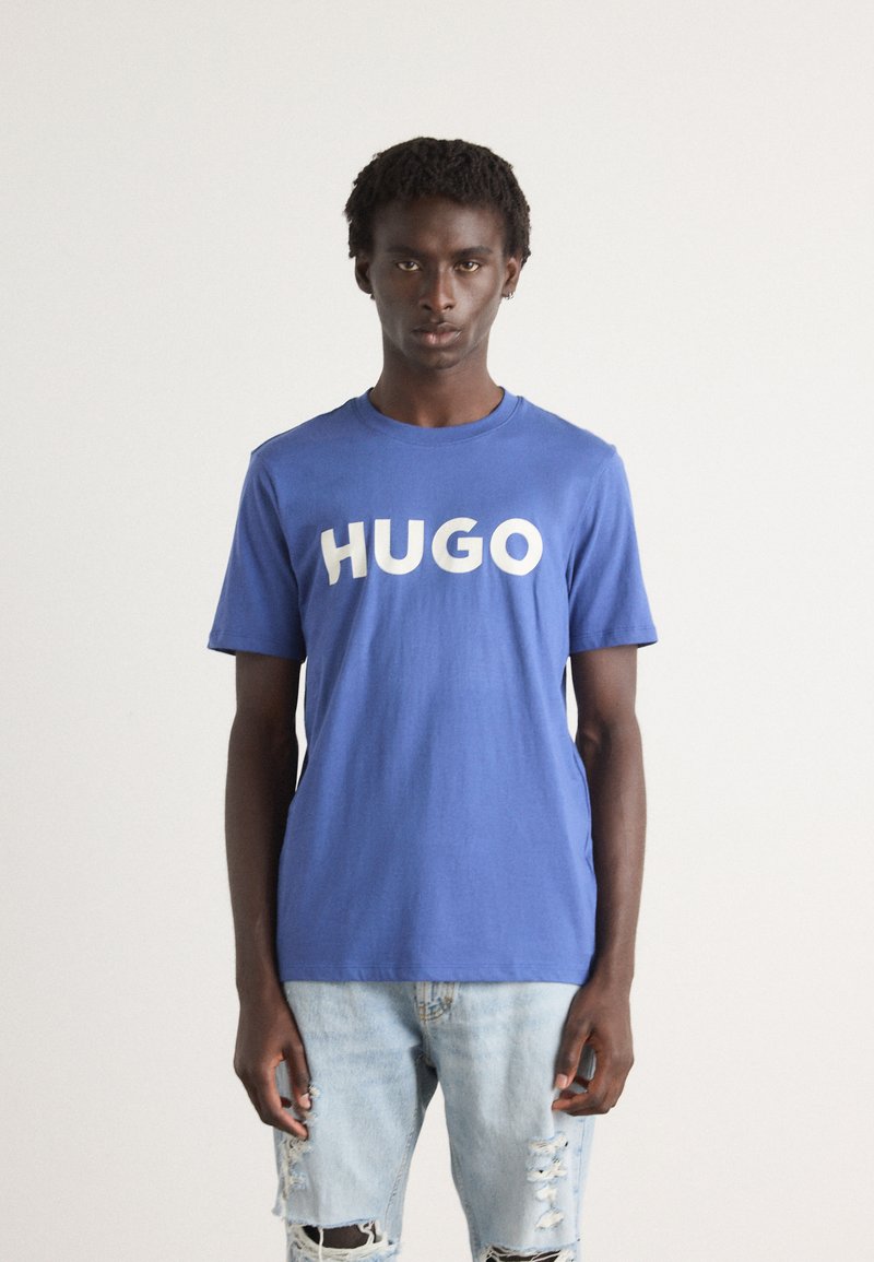 Kék pamut póló, nagy fehér "HUGO" felirattal az elején. Világoskék, kopottas farmernadrággal párosítva. Egyszerű, hétköznapi dizájn.
