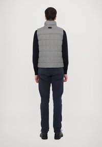 Gilet imbottito grigio a quadri con colletto alto, dotato di etichetta posteriore e abbinato a pantaloni blu navy e scarpe nere. Texture liscia.