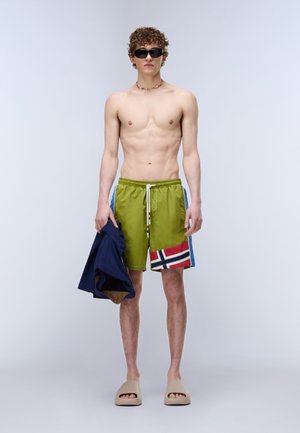 Ung mand med krøllet hår iført sorte solbriller, grønne badeshorts med flagdesign, holder en navyjakke og bærer beige klip-klappere.