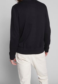Černý pletený svetr s ribbením podél výstřihu a manžet. Přiléhavý lem, nošený přes světle barevné kalhoty. Minimalistický design.