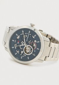 Ceas de mână automat din metal argintiu, cu cadran skeleton care arată roțile din interior, față neagră, marcaje orare albe și brățară din linkuri metalice.