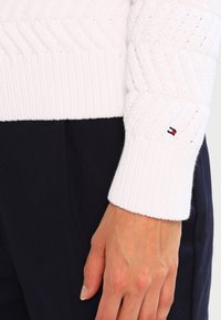 Vit stickad tröja med ett strukturerat sicksackmönster och ribbade ärmslut. Har en liten logotyp i rött, vitt och blått på ärmslutet.