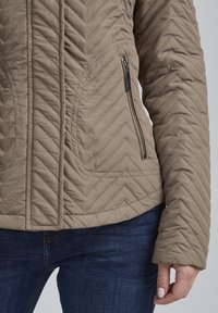 Karmelowa pikowana kurtka z wzorem w jodełkę, kieszenią na suwak i usztywnionymi rękawami, w zestawieniu z ciemnoniebieskimi jeansami.