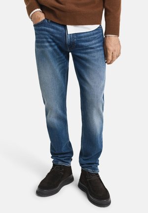 Blauwe denim jeans met een rechte pijp, lichte vervaging op de dijen, vijf zakken en een knoopsluiting, gecombineerd met bruine suède schoenen.