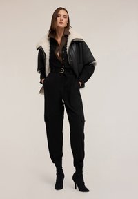 Zwarte faux leren jas met een witte shearling kraag, een bruine overhemd, zwarte joggers met riem, en zwarte enkellaarsjes met hak.