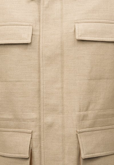 Κοντινό πλάνο μπεζ υφάσματος με τέσσερις τσέπες με καπάκι και ορατή ραφή σε ένα υφασμάτινο ένδυμα με υφή.