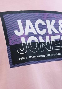 Lyserød T-shirt med et sort og lilla grafisk design med teksten "JACK & JONES." Designet inkluderer striber og tekst i fede hvide bogstaver.