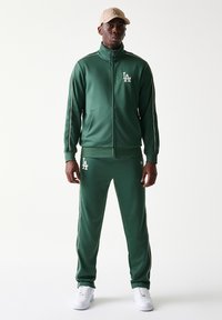 Πράσινη αθλητική φόρμα από λείο, ελαστικό ύφασμα με λευκές ρίγες. Διαθέτει μπουφάν με φερμουάρ, ελαστική μέση και κεντημένο λογότυπο.