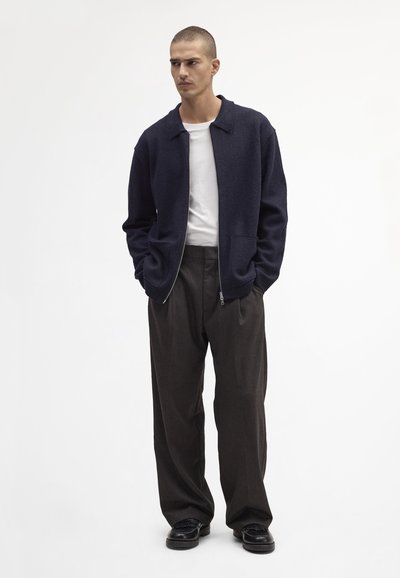 Marineblauwe wollen bomberjack met een kraag, een rits aan de voorkant en twee zakken, gecombineerd met donkere, wijde broekspijpen en zwarte schoenen.