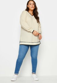 Top a maniche lunghe, color crema, con scollo a V e tessuto testurizzato, con accenti neri, abbinato a jeans skinny blu e sneaker bianche.