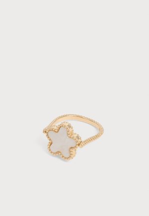 Bague en or avec un anneau perlée et un centre en forme d’étoile en nacre blanche, sur un fond clair uni.