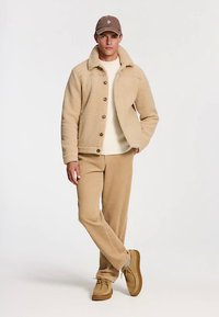 Bézs polár dzseki gombokkal, krémszínű inggel, bordázott anyagú bézs nadrággal és bézs cipővel párosítva. Barna sapka logóval.