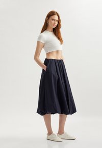 Gonna blu navy, lunga fino al ginocchio, con una cintura elastica e tasche laterali, abbinata a un top bianco corto e scarpe da ginnastica chiare. Tessuto morbido.