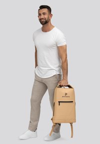 Mochila beige con un acabado mate, un mango superior, un bolsillo frontal con cremallera, logo y correas ajustables. Llevada por un hombre en atuendo casual.