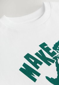 Weißes Baumwoll-T-Shirt mit grünem grafischen Text, der "MAKE" liest, und einem Designelement. Sichtbares geripptes Kragen-Detail. Weiche Textur und lässiger Stil.