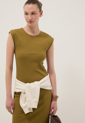 Frau trägt olivfarbenes ärmelloses Kleid mit weißem Sweatshirt, das an der Taille gebunden ist, hält eine braune Tasche vor schlichtem hellem Hintergrund.