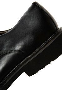 Zapato de cuero negro con una textura suave, tacón redondeado y una suela de goma con diseño. Presenta detalles de costura a lo largo del costado.