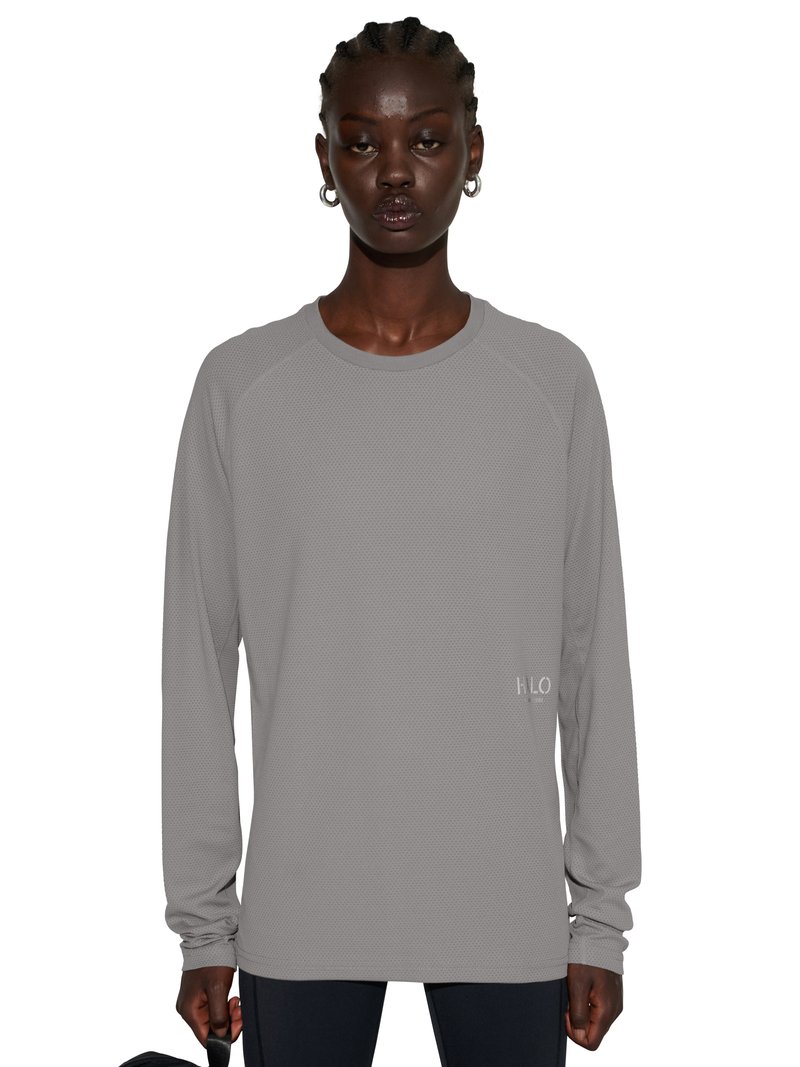 Modelka o ciemnej karnacji i splecionych włosach, ubrana w szarą teksturowaną koszulę z długim rękawem, trzymająca czarną rączkę torby.