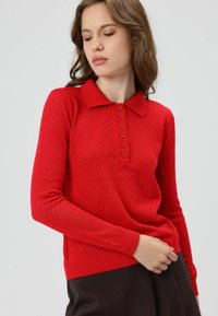 Rode gebreide trui met een ruitpatroon, geribde kraag en knoopsluiting. Lange mouwen met geribde boorden en zoom. Gladde textuur.