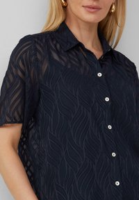 Marineblauwe blouse met knopen van doorzichtig materiaal en een textuur met golven; beschikt over een kraag en witte knopen.