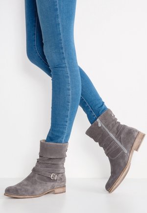 Jambes fines en jean bleu portant des bottines grises en daim souple avec fermeture éclair et boucle, debout devant un fond blanc.