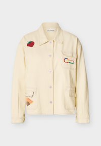 Κρεμ σακάκι με κουμπιά, με επιδιορθωτική ετικέτα φράουλας στον ώμο, πολύχρωμη καρφίτσα με χάντρες στην τσέπη του στήθους και επιδιορθωτική ετικέτα ανεμιστήρα στην κάτω τσέπη.