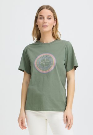 Kvinde iført en olivengrøn t-shirt med en rund pastel-farvet emblem og hvide bukser, stående mod en ensfarvet lys baggrund.